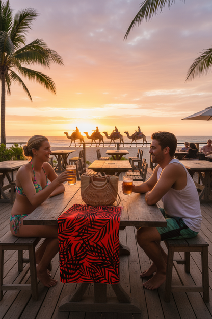 Two people sitting at a table on a beach at sunset, with palm trees and other beachgoers in the background.
